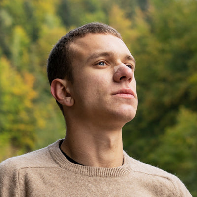 Junger Mann steht in der Natur und blickt nach oben, trägt ein transparentes Nasenpflaster auf der Nase.