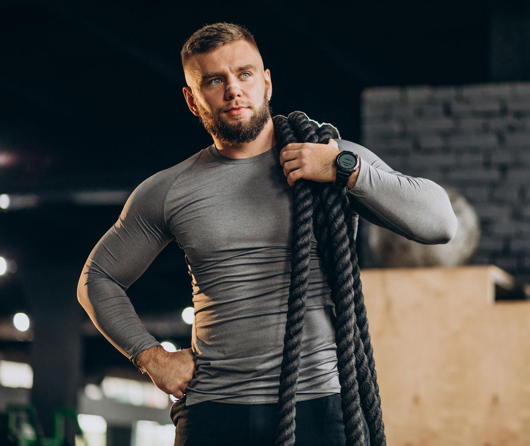Muskulöser Mann im grauen Sportshirt hält ein schweres Trainingsseil im Fitnessstudio – geeignet als Symbolbild für den Einsatz transparenter Nasenpflaster bei sportlichen Aktivitäten.