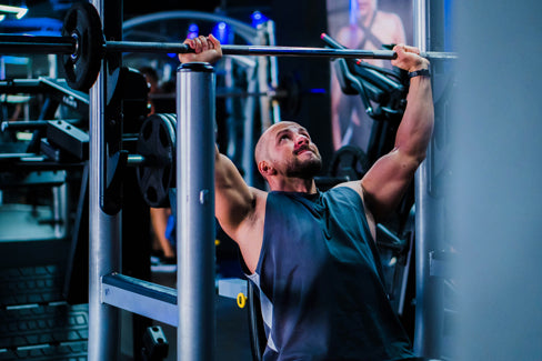 Sportler trainiert im Fitnessstudio an einer Langhantel im Sitzen, Blick nach oben gerichtet – geeignet als Symbolbild für den Einsatz transparenter Nasenpflaster bei sportlichen Aktivitäten.