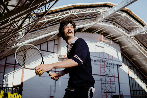 Ein junger Mann im Retro-Tennis-Outfit mit Nasenpflaster beim Tennisspielen unter einer überdachten Halle